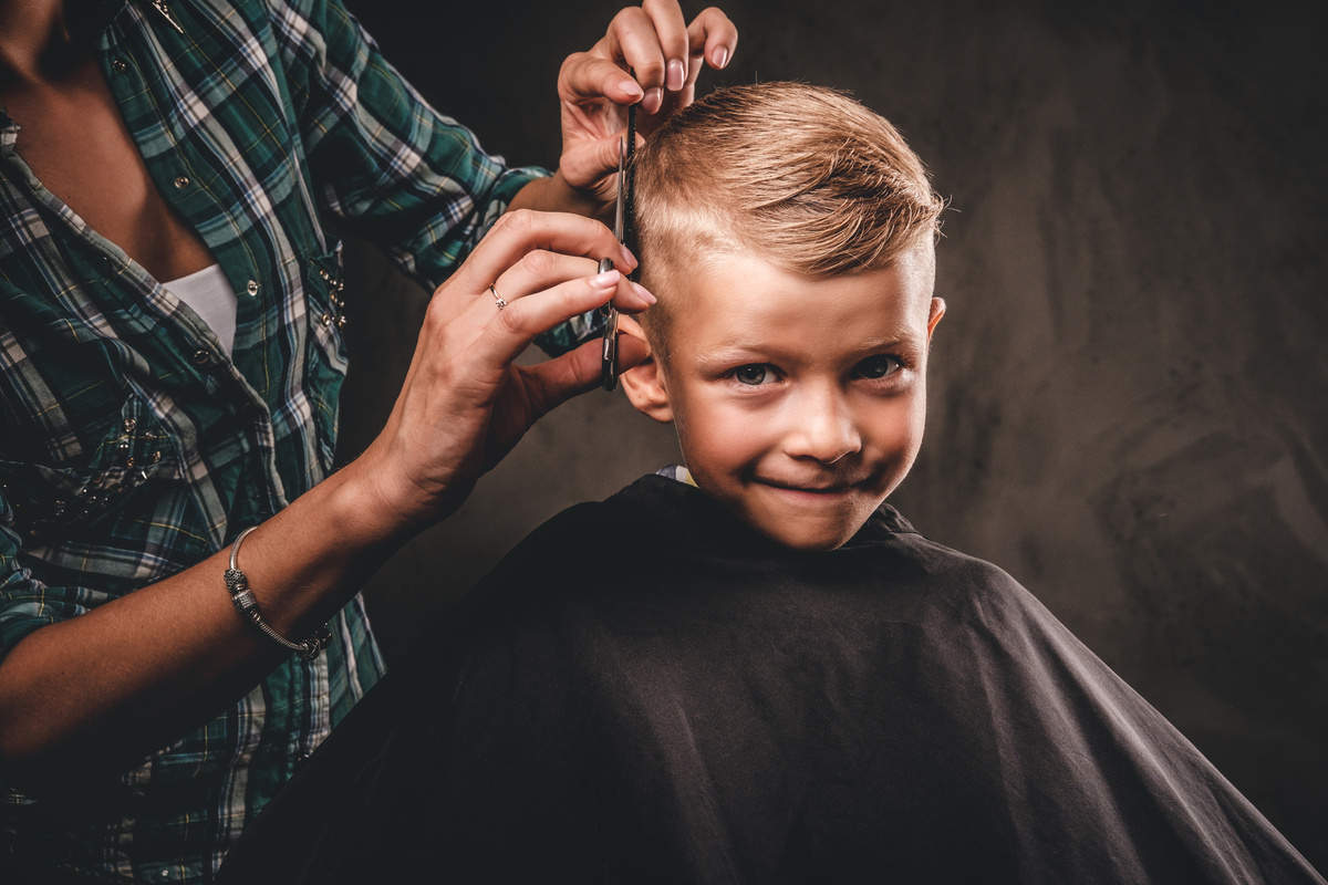 Children Hairdresser with Scissors Is Cutting Little Boy against